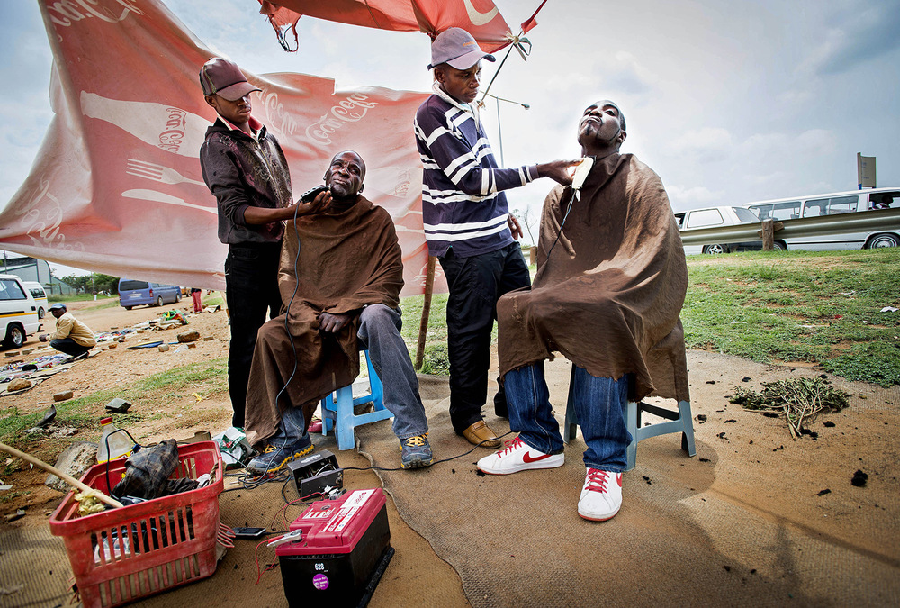 John T. Pedersen Photography - 2013 Barberers in Soweto Take-Away barbers are using a car battery, two razors and cooking oil. Julio Ernest (17) and Dick Mgwenya (30) manage to survive. By the noisy main road into Soweto, two men sit and shave for a couple of dollars each. Both hairdressers and customers are on alert. If the police show up, we just have to run. In the old days, we were kicked by the police and called donkey. Today we are free thanks to Father Madiba.